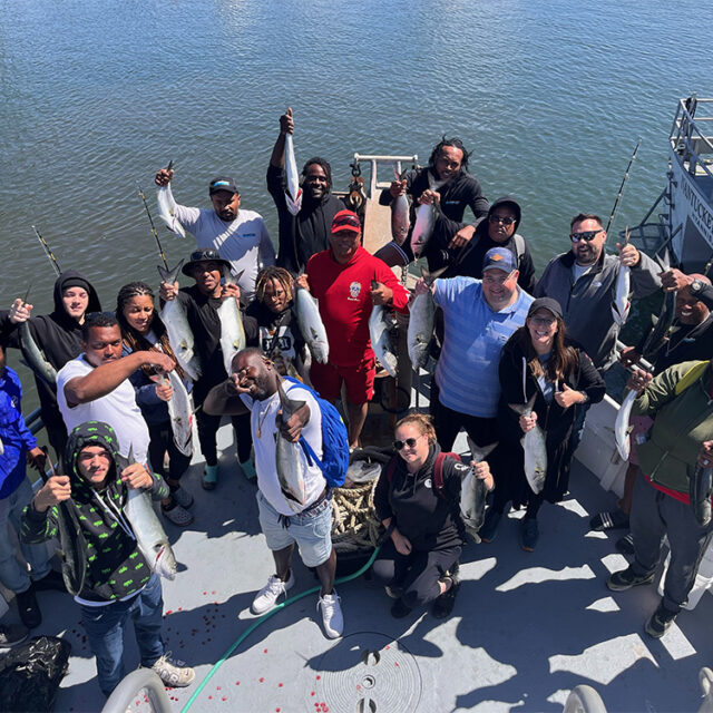 A group of people on a boat, displaying their caught fish while smiling and giving thumbs up. Water is visible in the background.