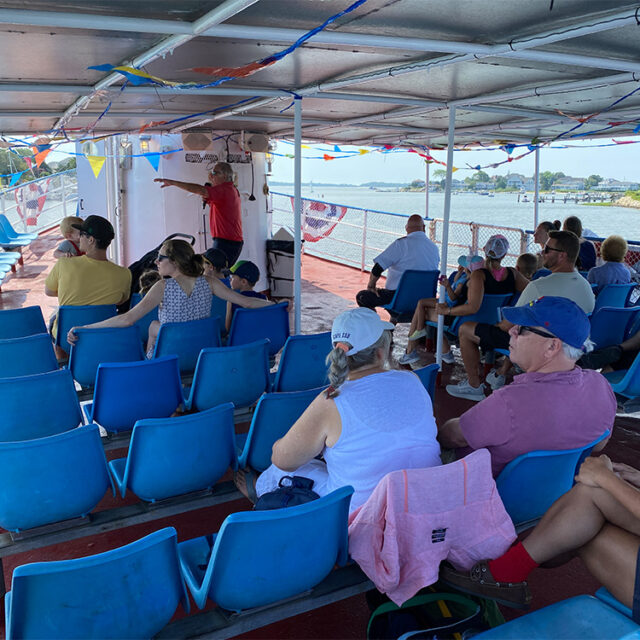 People are seated on blue chairs on a boat with a covered deck. A person is standing near the center speaking to the passengers. Decorated with colorful pennants. Land and water are visible in the background.