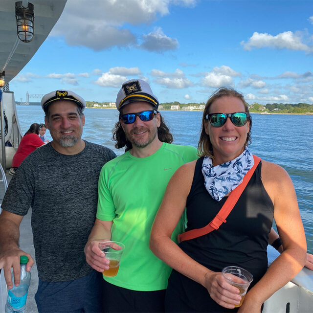 Three people wearing casual clothes and hats stand on a boat, smiling. Two of them are holding drinks. The background shows a body of water, a partly cloudy sky, and a shoreline with buildings.