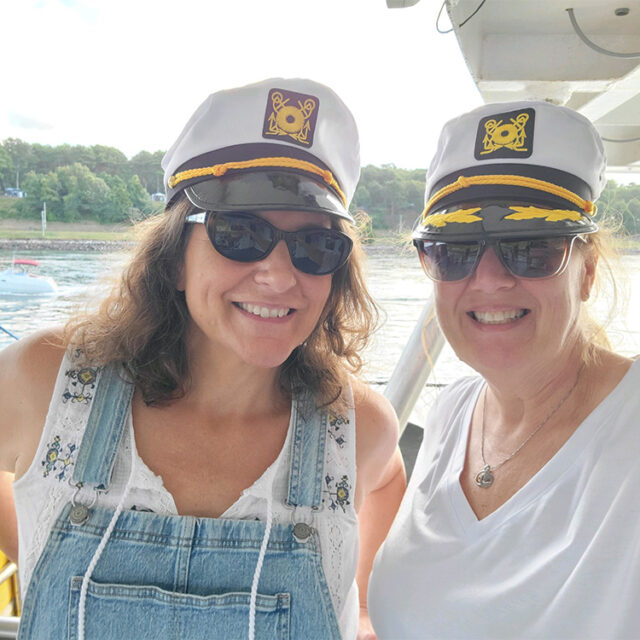 Two women wearing captain's hats and sunglasses smile while standing outdoors on a boat with water and greenery in the background.