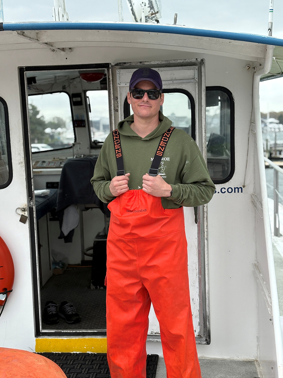young man standing on a Cape Cod fishing boat wearing sunglasses , sweatshirt and waders